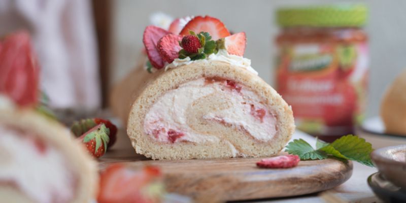 Eine Glutenfreie Biskuitrolle mit Erdbeercremefüllung liegt auf einem Holzbrett.