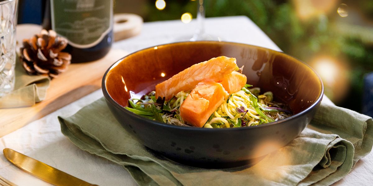 Eine Portion Lachs auf Gemüsepasta auf einem festlich gedecketem Tisch angerichtet, im Hintergrund sieht man verschwommen ein Weißweinglas und die Zweige eines Tannenbaums. Auf dem Teller ist Lachs auf Gemüsepasta angerichtet.